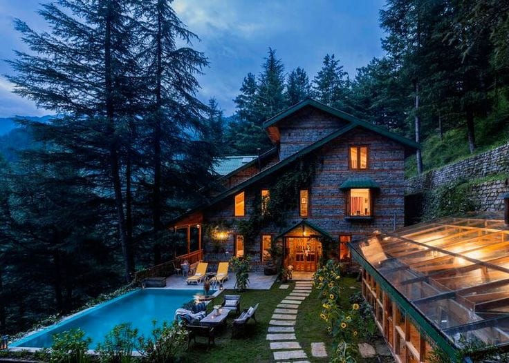 A warmly lit cottage and swimming pool beneath conifers