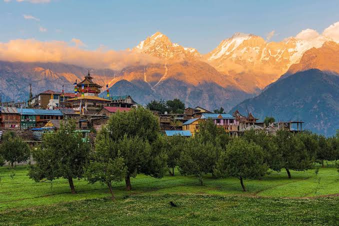 Village buildings below snow-capped peaks in warm evening light