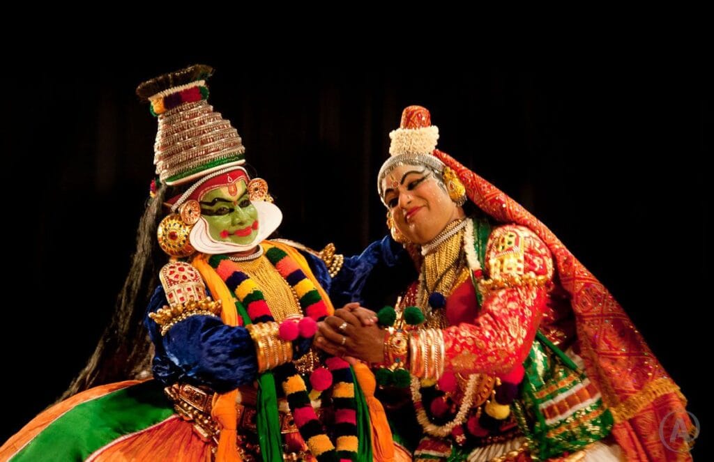 Two Kathakali performers in colorful costumes on a dark stage