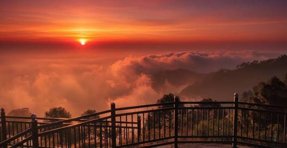 The sun setting above clouds beyond a hillside railing