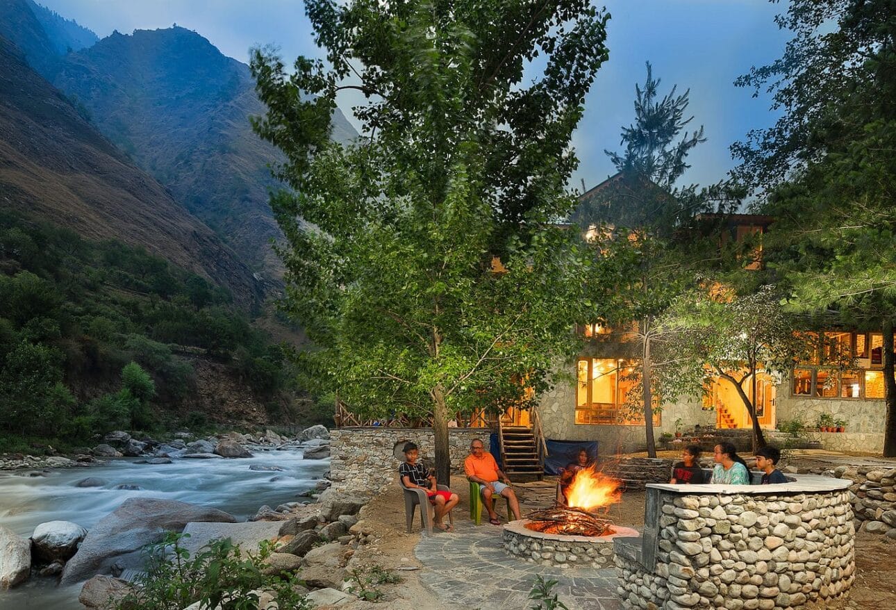 A campfire beside a river beneath leafy trees