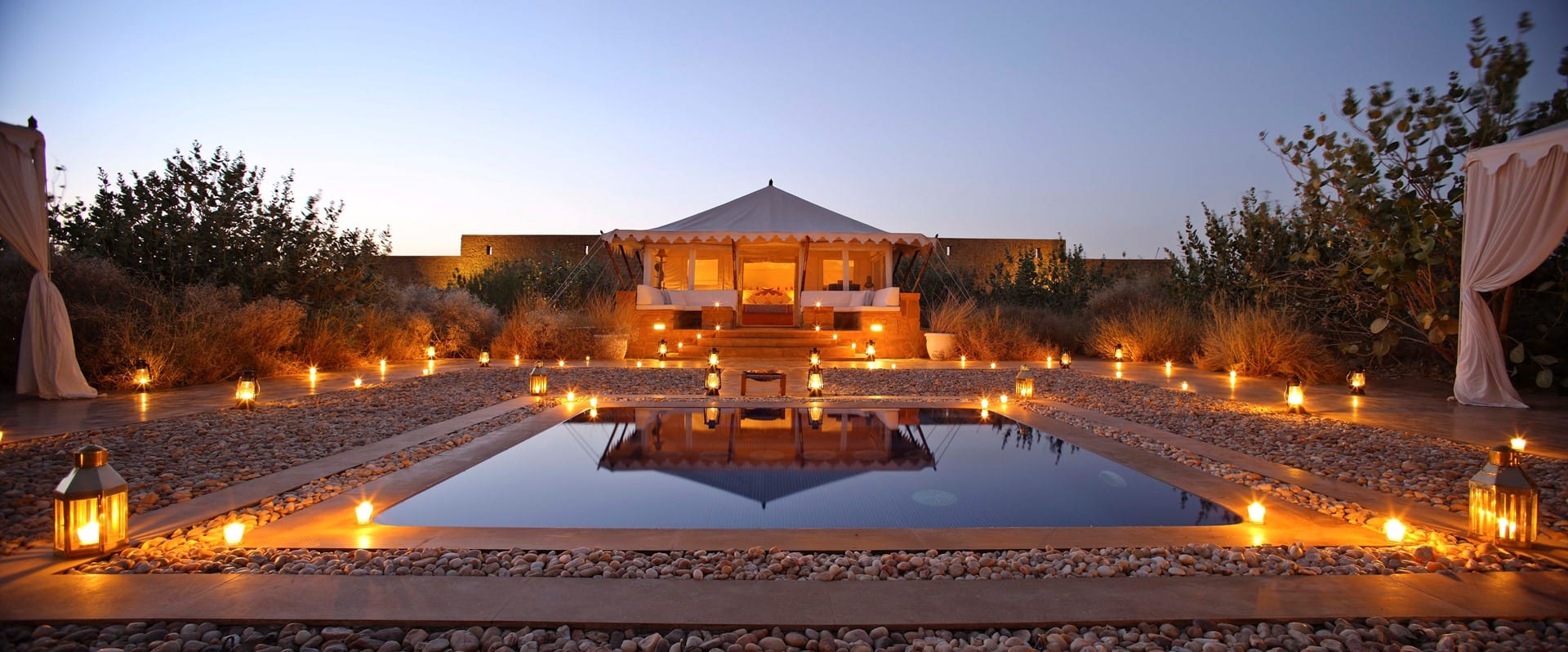 A lantern-lit courtyard and reflecting pool facing a tented lodge at dusk
